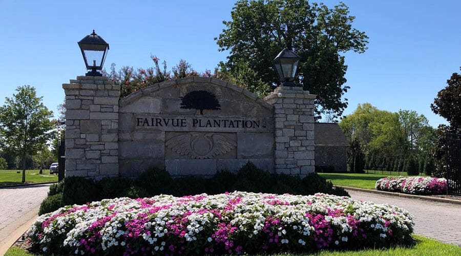 About Our Agency - Stone Entrance of Fairvue Plantation With Two Lamps and a Bed of White and Pink Flowers on a Sunny Day
